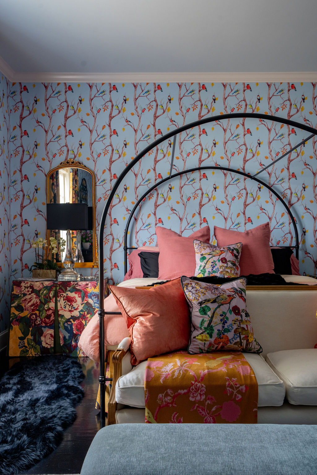 Bedroom with floral wallpaper, pink and patterned pillows, a canopy bed, floral side table, black lamp, gold mirror, and a black faux fur rug on a dark floor. Elegant window treatments frame the space beautifully, complementing the soft furnishings to create a cozy and inviting atmosphere.