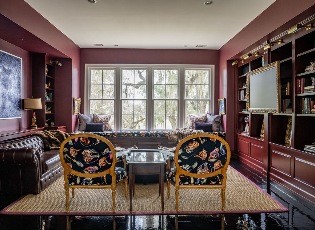 Library room with maroon walls, built-in bookshelves, a window seat, two floral chairs, a leather sofa, a rug, and thoughtfully chosen window treatments in front of large windows that flood the space with natural light. This inviting design blends comfort and elegance—perfect for curling up with a book or enjoying the serene savannah tones that inspire the room’s palette.