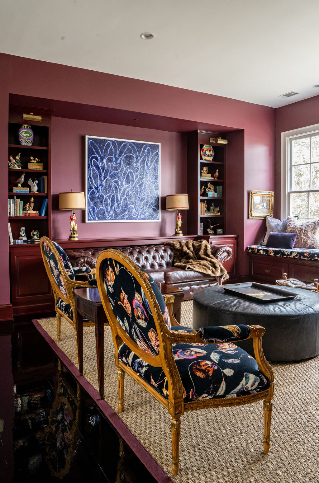 A living room with burgundy walls, patterned chairs, a leather sofa, built-in bookshelves, a large abstract painting, and a window seat is enhanced by elegant window treatments and thoughtful soft furnishings reminiscent of the relaxed charm of savannah style.