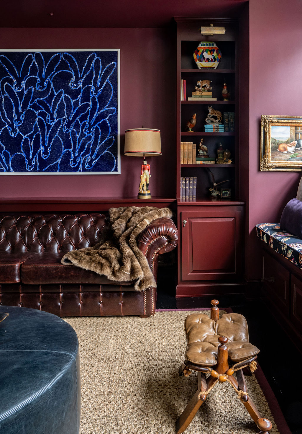 A maroon-walled living room with a leather sofa, fur throw, bookshelves, eclectic decor, thoughtful window treatments, a blue abstract artwork, and a brown leather footstool on a woven rug.