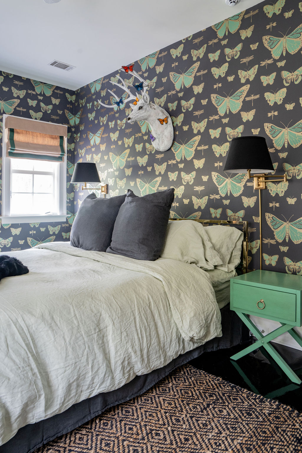 A bedroom with butterfly-patterned wallpaper, a bed with gray and white bedding, green nightstand, wall lamps, and a faux deer head wall mount above the headboard features thoughtful design touches such as custom window treatments and coordinating soft furnishings.
