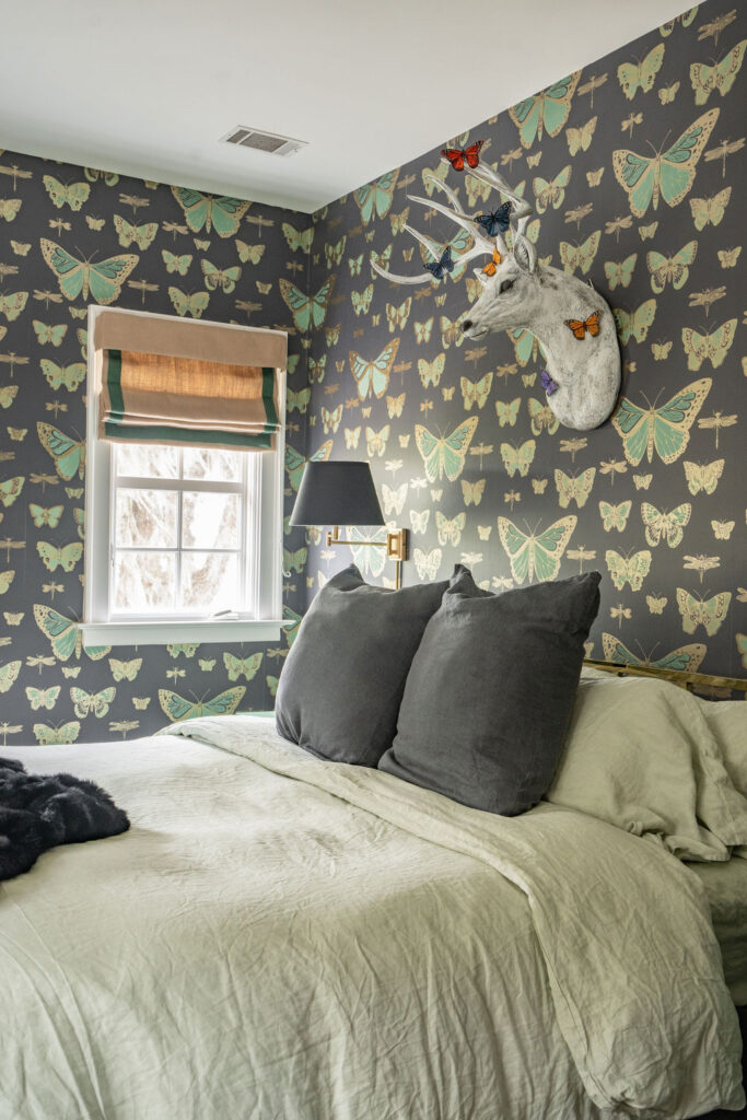 Bedroom with butterfly wallpaper, a mounted faux deer head, green bedding, two gray pillows, a window with a Roman shade as stylish window treatment, and a wall lamp—an inviting space where thoughtful design and soft furnishings create an enchanting atmosphere.