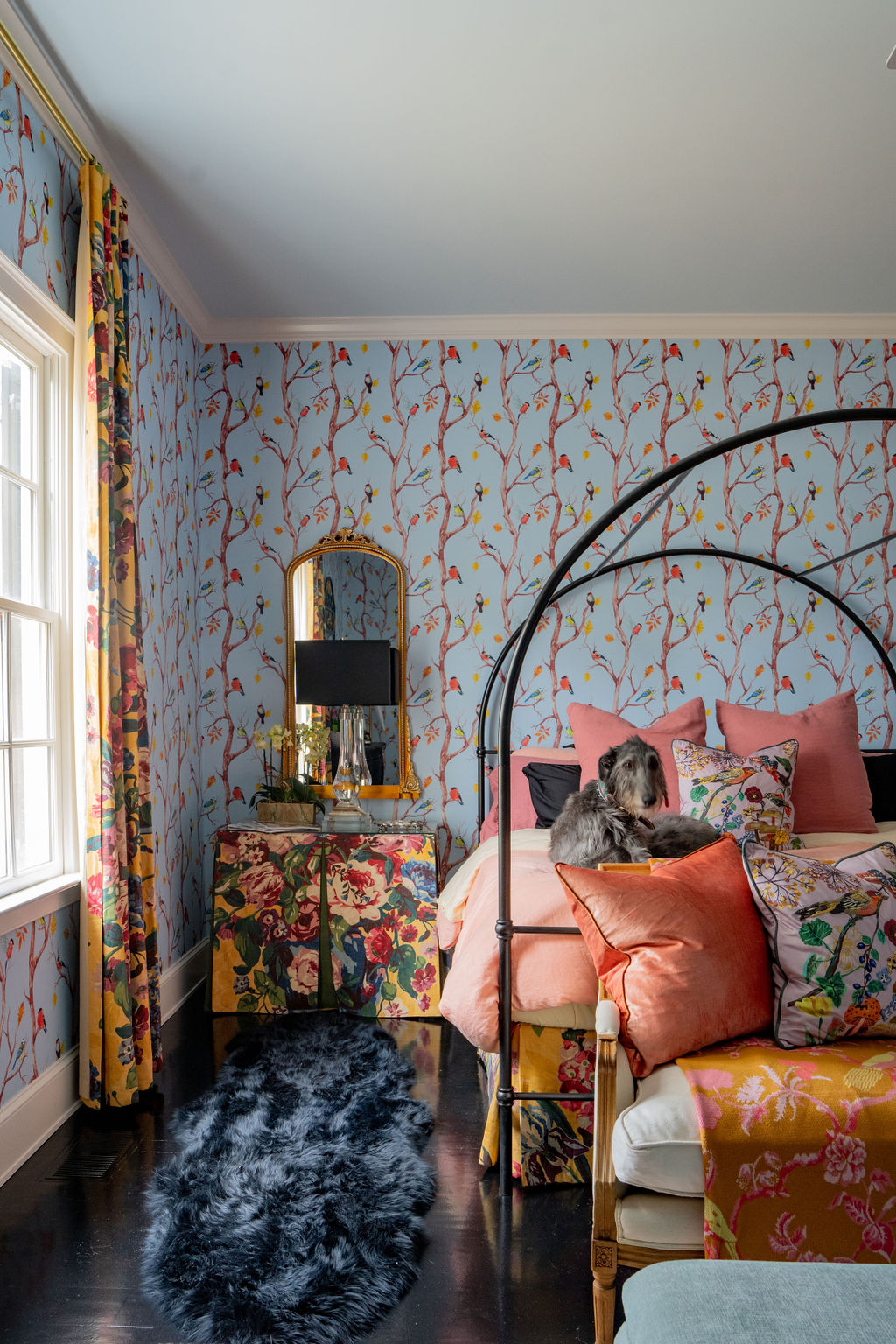 Colorful bedroom with floral wallpaper, mixed patterned textiles, a black canopy bed, decorative pillows, and thoughtfully selected soft furnishings that elevate the design. A nightstand with a tray sits nearby, while a dog lounges comfortably on the bed, completing this vibrant savannah-inspired retreat.