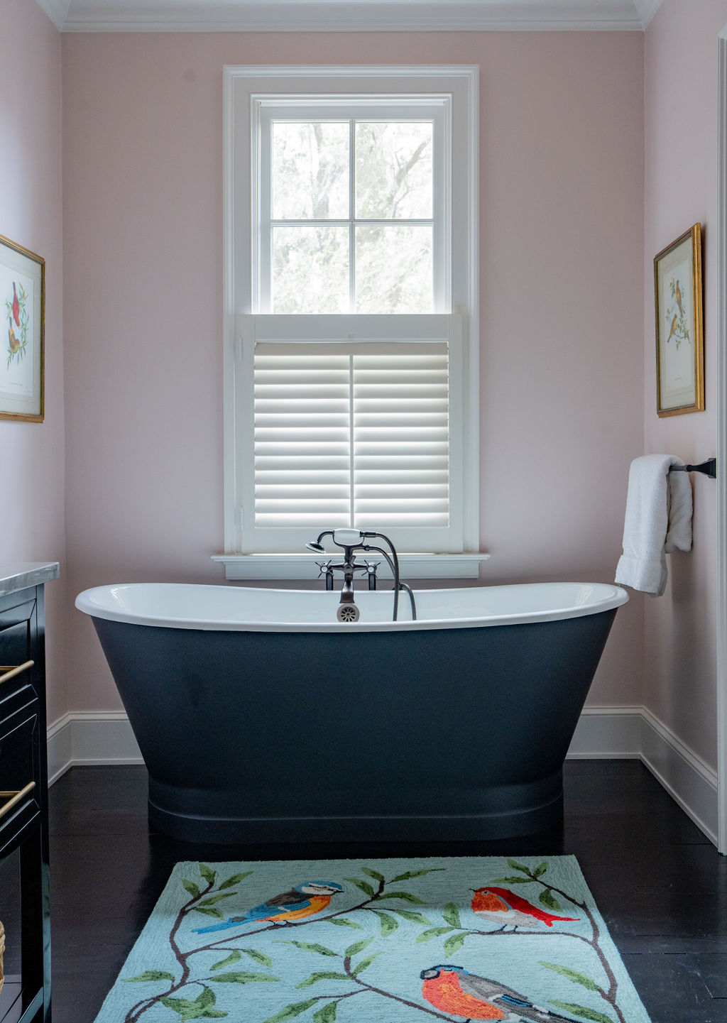 A black freestanding bathtub sits under a window with white shutters and custom window treatments in a pink-walled bathroom, with a colorful bird-themed rug and coordinating soft furnishings on the dark floor.