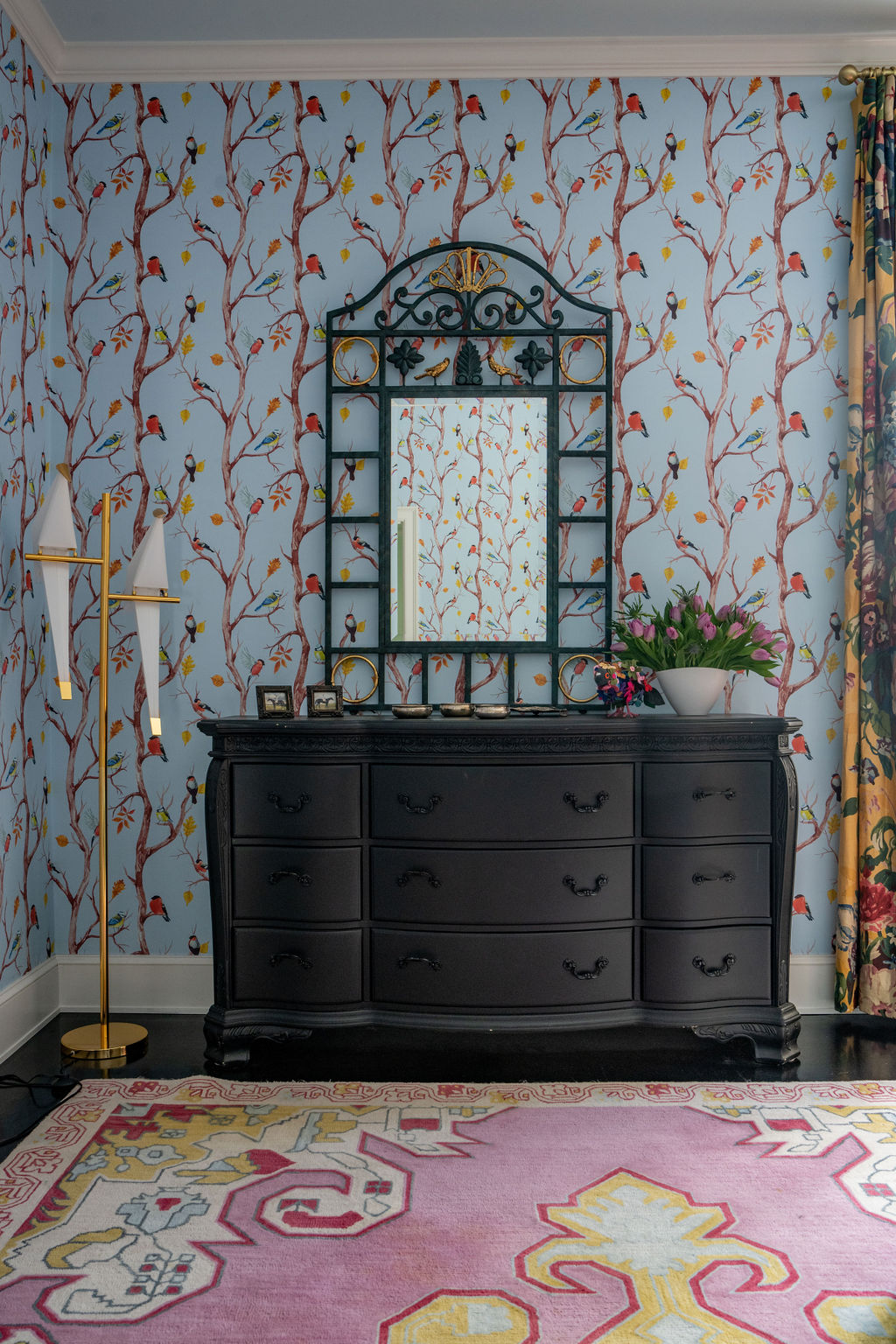 Room with floral wallpaper, black dresser, ornate mirror, gold floor lamp, large pink rug, and floral curtains; vase with flowers and framed photos on dresser—thoughtfully layered soft furnishings and custom window treatments complete the charming design.