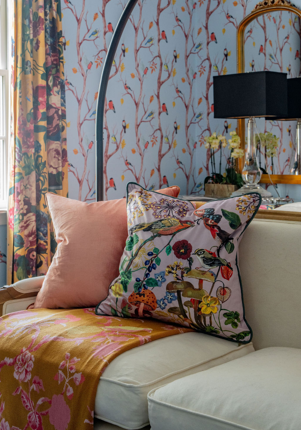 A white sofa with pink and floral pillows, a yellow patterned throw, and floral wallpaper with trees and birds brings savannah-inspired charm to this colorful living room, where soft furnishings and thoughtful design create a welcoming atmosphere.